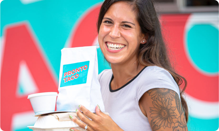 Woman with a big smile looking at the camera holding a taco box and bag of chips in front of a food truck