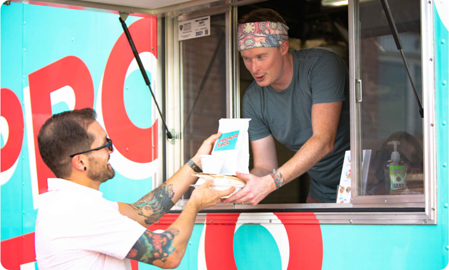 Food truck employee with a bandana handing a taco box order to customer who is taking the order from the employee