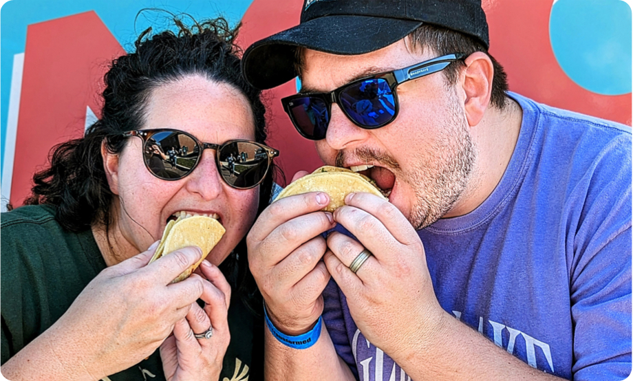 Couple wearing sunglasses eating tacos with food truck behind them