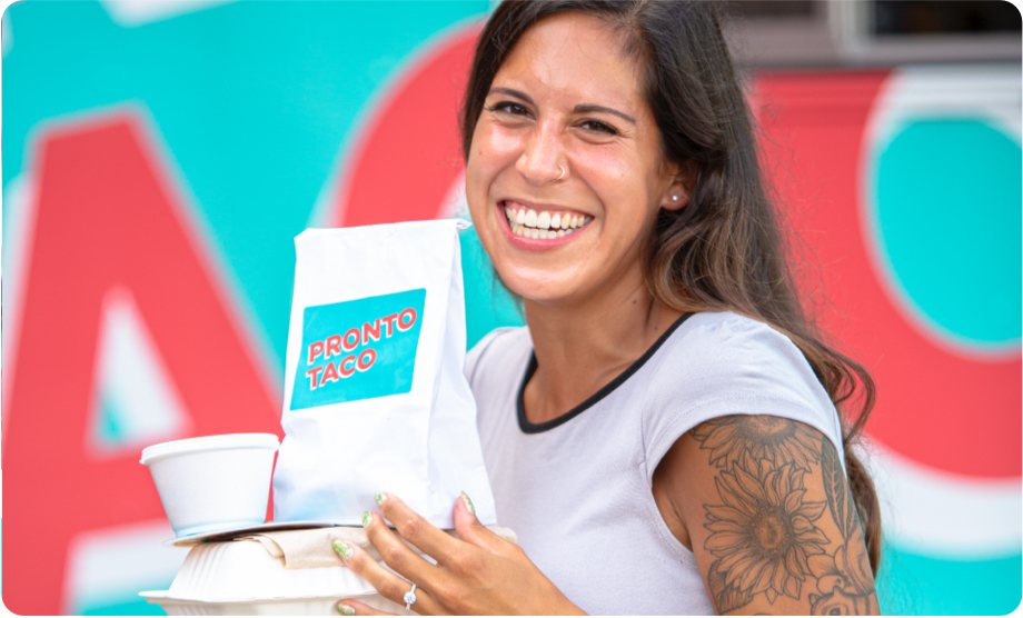 White woman with long dark brown hair, a nose ring, and flower tattoos smiling at the camera while carrying food containers
