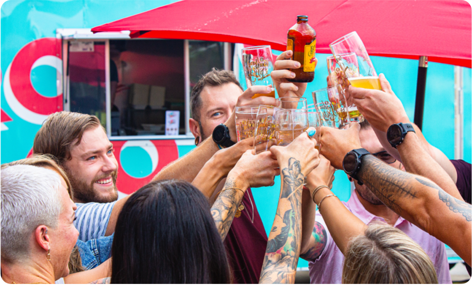 Group of people in front of a food truck celebrating a cheers with glasses of beer in their hands