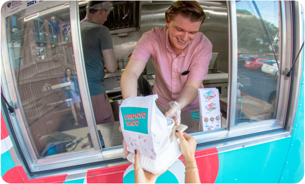 Fish eye photo of smiling food truck employee handing food to unknown person (only hands are shown)
