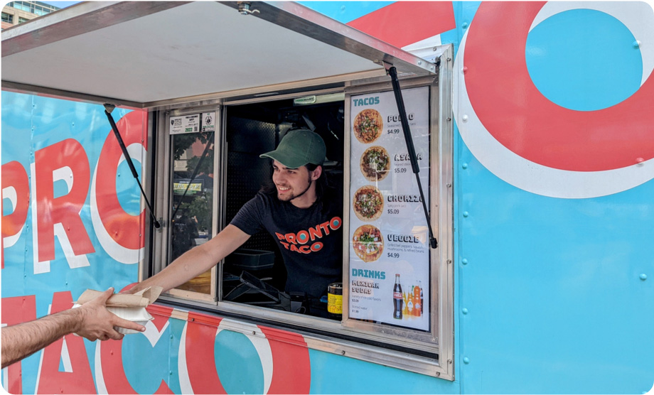 Food truck employee leaning out of the food truck window handing a taco box order to a customer