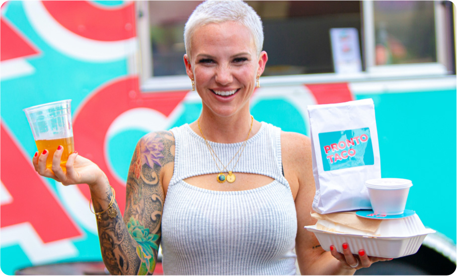Short haired woman in front of a food truck holding a box of tacos and bag of chips in one hand and a glass of beer in the other hand