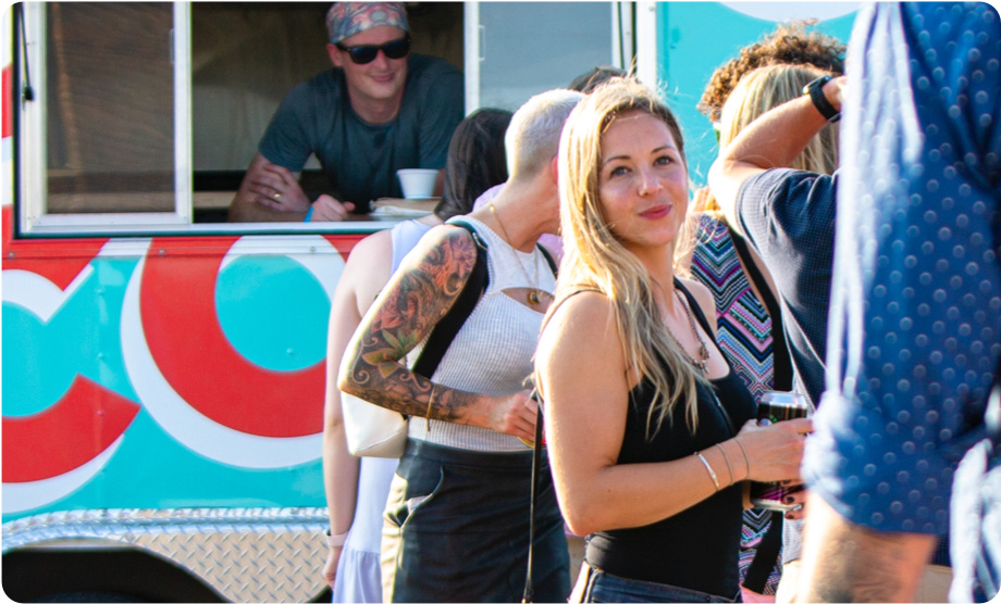 Woman in line with other people at food truck window smiling at her boyfriend whose arm is in the foreground