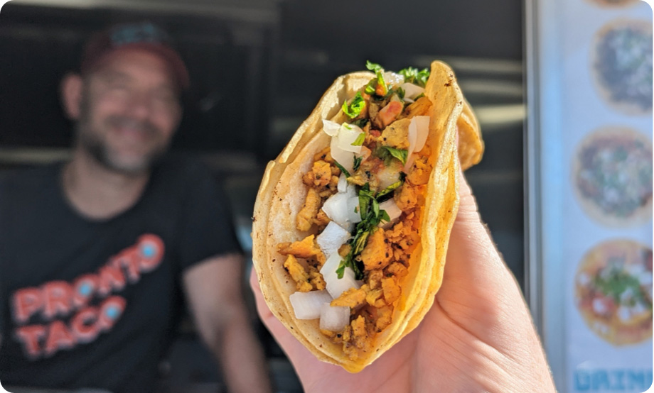 Closeup of hand holding taco with Pronto Taco employee in food truck in background