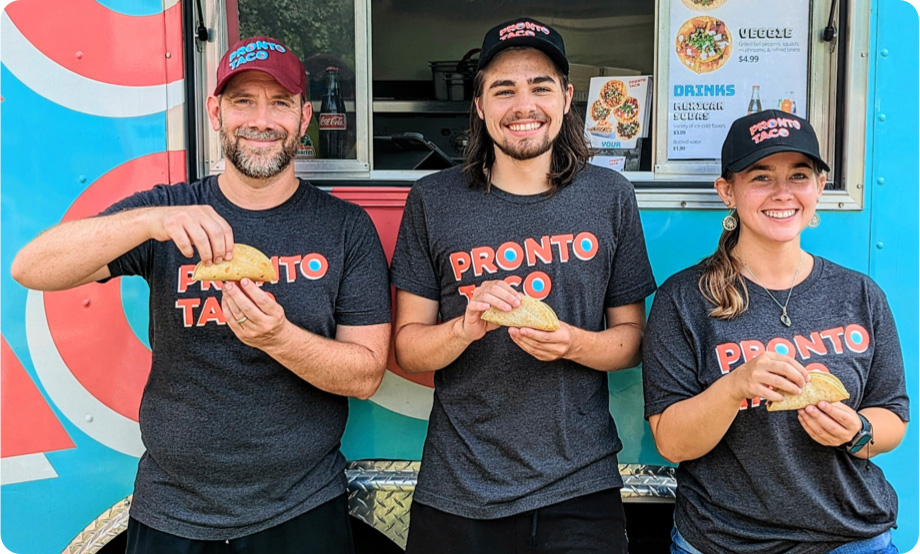 Team of three food truck employees holding tacos and smiling at the camera