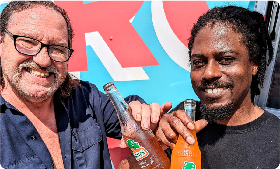 Two men smiling with Mexican sodas: a white man with shoulder length hair, a beard and glasses, and a black man with a mustache and goatee